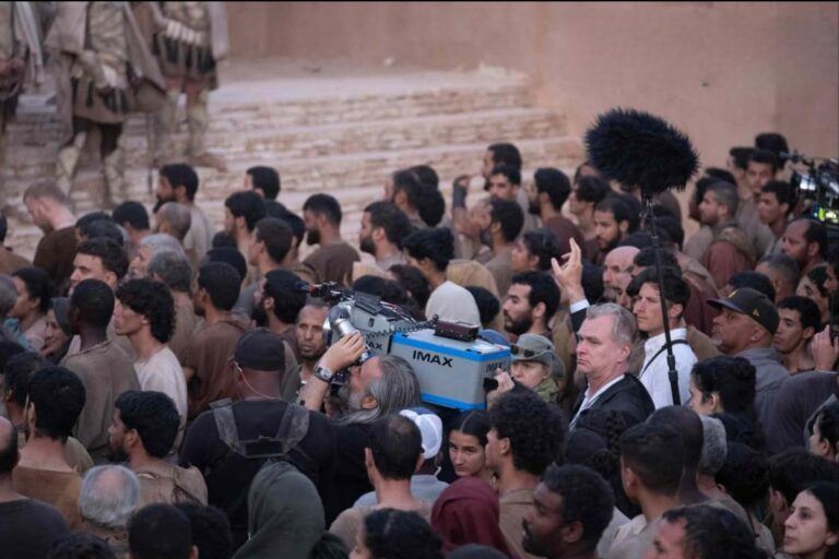A large crowd in period-style costumes surrounds a film crew operating an IMAX camera on set, capturing an epic scene from The Odyssey
