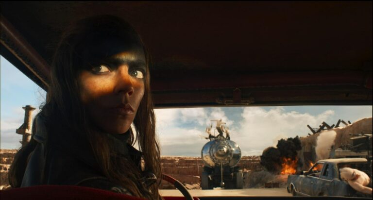 A determined woman with dark war paint sits inside a vehicle, staring intensely ahead as explosions and chaos erupt in a desert wasteland in Furiosa: A Mad Max Saga.