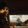 A determined woman with dark war paint sits inside a vehicle, staring intensely ahead as explosions and chaos erupt in a desert wasteland in Furiosa: A Mad Max Saga.